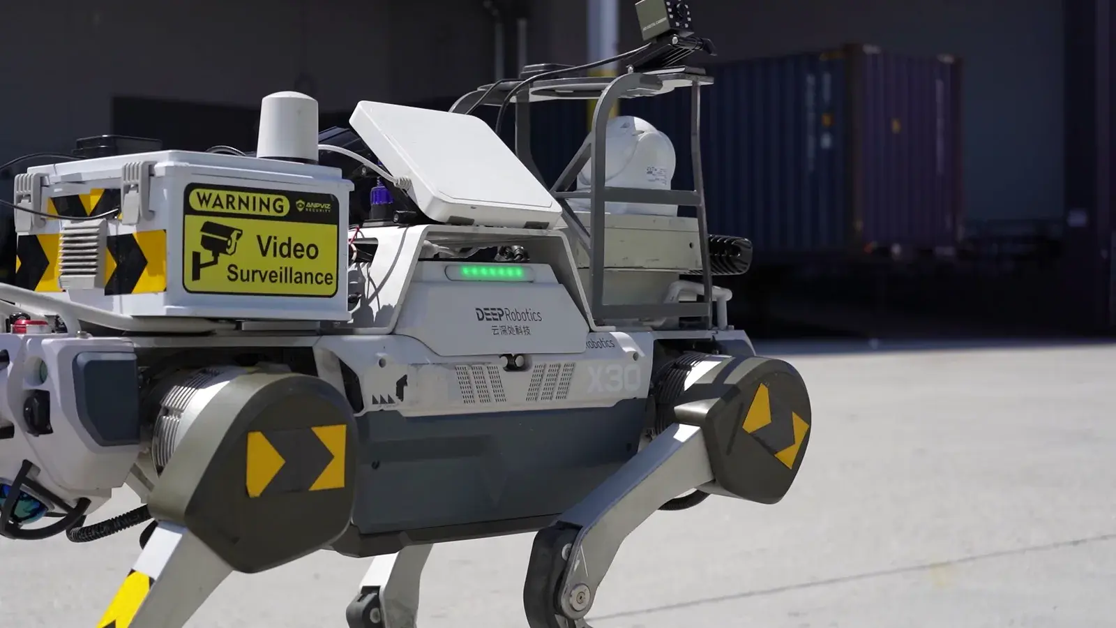 A RobotX quadruped inspection robot with a surveillance payload walking past shipping containers at Shiplink's logistics yard.