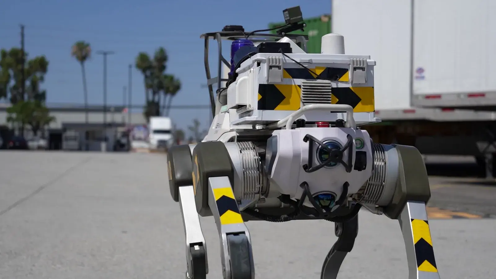 The quadruped inspection robot walking across the open yard with parked trailers behind it.